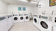 This shared laundry facility is a practical, efficiently designed space that accommodates a full suite of commercial-grade laundry equipment. The room is finished with light-toned walls and durable tile flooring, creating a clean and bright environment enhanced by overhead fluorescent lighting that ensures excellent visibility throughout.

Multiple front-loading washing machines are arranged in organized rows along the walls, accompanied by a corresponding bank of top-loading dryers, providing ample capacity for simultaneous use. The machines feature digital control panels for ease of operation, and instructional posters displayed on the walls offer guidance on proper usage and detergent recommendations.

A dedicated folding and sorting area is positioned along one wall, featuring a built-in countertop with a checkered surface, paired with a chair for added convenience. A self-serve electronic payment kiosk is mounted near the entrance, streamlining the payment process for users. Additional wall-mounted notices and information boards keep users informed of facility rules and updates.

A fire extinguisher is visibly mounted near the entry point, reflecting attention to safety standards. The layout is thoughtfully organized to allow comfortable movement between machines, making this laundry facility a functional and user-friendly shared amenity.