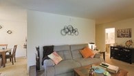 This single-story space features a thoughtfully arranged living area that serves as the central gathering point. The living room is finished with smooth, light-colored walls that create a bright and airy atmosphere. A large, comfortable gray sofa anchors the seating area, accented with warm orange and floral throw pillows that add pops of color against the neutral palette. A wooden coffee table sits in front of the sofa, providing both function and a natural design element. Decorative touches include a striking metal wall sculpture composed of overlapping circular rings, adding an artistic focal point to the room. A tall, slender decorative cat figurine stands beside the sofa, contributing to the eclectic character of the space. The flooring transitions from a low-pile carpet in the living area to what appears to be a hard surface in the adjoining room, visible in the background. The layout offers an open sightline into a secondary area, suggesting a flowing, connected floor plan. Overall, the space presents a casual yet stylish ambiance, blending comfort with decorative personality in a well-maintained, single-level setting.