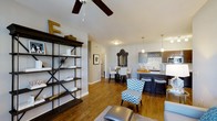 This single-floor residence showcases an open-concept layout that connects the living, dining, and kitchen areas into one cohesive, functional space. Warm hardwood flooring runs throughout, lending a sense of continuity and warmth across the entire floor plan.

The living area features a ceiling fan overhead and is furnished with eclectic, eye-catching pieces, including a chevron-patterned accent chair and a decorative open metal shelving unit displaying curated décor items. A textured area rug anchors the seating zone.

The dining area sits centrally within the space, anchored by a dark dining table with white upholstered chairs and accented by a bold ornate mirror mounted on the wall, adding a dramatic focal point.

The kitchen is positioned along one side, featuring dark cabinetry, a subway tile backsplash, stainless steel appliances, and pendant lighting above a breakfast bar with bar-height seating. The bar counter provides additional casual dining options and reinforces the open connection between the kitchen and living areas.

A hallway visible in the background suggests additional private rooms beyond the main living space, completing this well-appointed, contemporary layout.