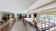 The Upper Clubhouse at Woodland Village in San Marcos is a well-appointed community facility designed to offer residents a variety of social and recreational spaces. The layout spans a single floor and is organized into several distinct yet open areas, connected by wide carpeted corridors lined with warm-toned, floral-patterned carpet throughout.

A central hallway features elegant wood railings overlooking the lower level, with a decorative chandelier and large arched windows that invite abundant natural light and views of the surrounding greenery. The open design and coffered ceilings lend the space a refined, welcoming ambiance.

The game room area is furnished with multiple round card tables and cushioned leather chairs, accommodating group play and socializing. Glass-paneled doors lead to an adjacent billiards room, while decorative display cases add character to the space.

A dining area features a large rectangular table set beneath an arched window framed by lush indoor plants, with a built-in credenza providing functional storage. The room is bright and inviting, ideal for shared meals or small gatherings.

A library and activity nook offers bookshelves, a high-top table, and comfortable seating, creating a quiet retreat for reading or games. Throughout the clubhouse, recessed lighting, structured ceiling details, and neutral wall tones contribute to a cohesive, comfortable atmosphere suited for community engagement.