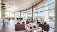 Located at 3242 Peachtree Rd NE in Atlanta's prestigious Buckhead district, this upscale lounge space occupies a single floor and offers a seamlessly designed environment built for social gathering and relaxation. The most striking architectural feature is the sweeping curved curtain wall of floor-to-ceiling glass that wraps around the perimeter, flooding the interior with abundant natural light and framing breathtaking panoramic views of the Atlanta skyline and surrounding cityscape.

The open-plan living area is thoughtfully organized into distinct zones. A lounge area is anchored by richly upholstered leather seating arrangements atop layered area rugs set over large-format wood-look tile flooring, creating a warm yet refined atmosphere. Slim structural columns punctuate the curved glass wall without interrupting the views.

Adjacent to the lounge, a full-length bar with white countertops and sleek backless barstools commands attention, accented by a dramatic textured feature wall and stylish pendant lighting overhead. Curated contemporary artwork adorns the walls throughout, contributing to the space's polished, gallery-like aesthetic.

A dining area with round tables and modern chairs sits within the curved glass bay, offering an ideal setting for meals with unobstructed city views. Recessed lighting complements the natural light, ensuring the space is equally inviting after dark. The overall ambiance is sophisticated, modern, and distinctly urban.