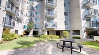 This outdoor communal space is nestled within the interior courtyard of a multi-story residential building complex. The area is thoughtfully designed to offer residents a comfortable and inviting outdoor environment, combining hardscape and softscape elements in a cohesive layout.

At the heart of the space is a generously sized paved patio constructed with square stone tiles, providing a level and durable surface ideal for outdoor gatherings. A metal picnic table and bench set is positioned on the patio, offering residents a dedicated area for outdoor dining or socializing. The patio transitions seamlessly into well-kept green lawns that extend throughout the courtyard, creating an open and airy feel.

The landscaping is a standout feature of this space, with a variety of mature trees, ornamental shrubs, and seasonal plantings arranged in carefully maintained garden beds. Decorative river rock borders and raised stone retaining walls add structure and texture to the planting areas. Wrought iron fencing encloses portions of the perimeter, providing a sense of security while maintaining visual openness to the surrounding greenery.

The multi-story building facades, featuring private balconies with black metal railings, frame the courtyard on multiple sides. Ground-level access points and covered entry areas connect the outdoor space to the building's interior. The overall ambiance is calm, green, and well-organized, making this courtyard a practical and pleasant shared outdoor amenity.