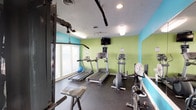 This fitness center is a purpose-built exercise space designed to support a well-rounded workout routine. The room is immediately recognizable by its energetic color scheme, featuring bold lime green and teal accent walls that create a lively, motivating atmosphere. Durable rubber flooring covers the entire space, providing both safety and functionality for a variety of exercises.

The cardio section is well-stocked, housing multiple treadmills and an elliptical trainer, allowing several users to engage in aerobic workouts simultaneously. A wall-mounted flat-screen television is positioned for easy viewing during cardio sessions, and a tower speaker adds audio options for an enhanced workout experience. A ceiling-mounted pull-up bar and a multi-station cable strength machine provide options for resistance and strength training, while a spin bike, rowing machine, kettlebells, and free weights round out the equipment offerings.

Large windows along one wall flood the space with natural light and offer views of the surrounding outdoor landscape, contributing to an open and airy feel. Full-length wall mirrors expand the visual depth of the room and allow users to monitor their form during exercise. Recessed and panel lighting ensures the space remains well-lit throughout. Access doors on opposing walls provide convenient entry and exit points to the facility.
