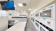 Located at 226 Dorchester Dr in East Windsor, NJ, this commercial laundromat offers a well-organized, single-floor facility designed for efficiency and customer comfort. The interior is finished in clean white walls with a durable speckled terrazzo-style floor, creating a bright and sanitary environment throughout.

The main laundry area is lined with rows of top-load and front-load washing machines, including stacked dryer units from commercial brands such as Hercules. Machines are clearly numbered for easy identification and are arranged along both sides of the room, maximizing the available floor space and allowing for smooth customer flow. Folding tables are conveniently positioned throughout the facility to assist with garment care after cycles are complete.

A dedicated waiting area is situated at the rear of the space, featuring large windows fitted with white horizontal blinds that allow natural light to fill the room. A wooden bench provides comfortable seating, accompanied by a table and chair for customer use. A wall-mounted television and posted laundry instruction guides add to the practical amenities available.

The facility also includes a wall-mounted HVAC unit, overhead recessed panel lighting, and clearly marked exit signage, reflecting attention to safety and comfort standards. A utility cart is available for customers to transport laundry with ease. The space is neatly maintained and thoughtfully laid out to serve a high volume of users efficiently.