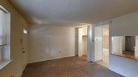 This single-floor space features a straightforward, functional layout finished in neutral tones. The main room is carpeted in a warm beige, complemented by smooth, light-colored walls that create a clean, understated atmosphere. A textured popcorn ceiling adds a classic touch overhead, while a floor vent indicates a central HVAC system for climate control.

A notable feature is the walk-in closet, accessible through a bifold door, offering organized storage with a visible hanging rod and shelving system. Adjacent to the closet, a sliding door leads to a connected room, suggesting a practical flow between spaces. The glimpse into the neighboring area reveals a tiled or hard-surface floor, contrasting with the carpeted main room and hinting at a bathroom or utility space.

The overall design is simple and utilitarian, prioritizing functionality over ornamental detail. Natural light appears limited based on the image, with interior lighting providing illumination. The space presents a move-in-ready condition with clean walls and well-maintained flooring throughout.