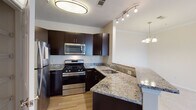 This single-floor residence offers a thoughtfully designed layout with a seamless flow between its key living areas. The kitchen is a standout feature, showcasing rich dark cabinetry, light granite countertops, stainless steel appliances including a gas range, built-in microwave, and refrigerator, all set atop warm hardwood-style flooring. A peninsula counter extends the workspace while creating a natural transition into the adjacent dining area, which is accented by an elegant chandelier overhead and track lighting nearby.

The living area is generously proportioned, bathed in natural light streaming through a full-height sliding glass door fitted with vertical blinds that open to an outdoor space. Soft, neutral-toned carpeting runs throughout the living, dining, and bedroom areas, contributing to a warm and comfortable ambiance. Crown molding adds a refined architectural detail to the main living spaces.

The bedroom is a quiet, well-lit retreat featuring a ceiling fan with light fixture and a window with blinds that allow for natural light and privacy. A walk-in closet with built-in shelving provides ample storage. The bathroom includes a full soaking tub and shower combination with clean white tile surround accented by a neutral tile border, offering a crisp, functional finish.
