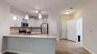 Located at 7108 Woods Walk in Villa Rica, Georgia, this single-floor residence offers a thoughtfully designed open layout that promotes a comfortable and functional living experience. The kitchen is a standout feature, showcasing white shaker-style cabinetry with glass-front upper panels, granite countertops, a tiled backsplash, and stainless steel appliances including a range, microwave, and refrigerator. A peninsula with pendant lighting provides additional prep space and serves as a natural divider between the kitchen and the main living area. The living space is bright and open, with wood-look flooring underfoot and French doors that invite natural light and provide outdoor access. Bedrooms are carpeted for comfort, with large windows offering views of the surrounding greenery. At least one bedroom includes a walk-in closet with built-in shelving. The primary bathroom features a granite-topped vanity, wood-look tile flooring, a glass-enclosed shower, and accessibility grab bars. A secondary bathroom mirrors the same refined aesthetic with a granite vanity and a full tub-shower combination. Throughout the home, neutral tones, clean lines, and consistent finishes create a cohesive and welcoming atmosphere.