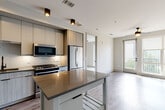 This contemporary single-floor residence showcases a thoughtfully designed open-concept layout that seamlessly connects the kitchen and living areas. The kitchen is a standout feature, equipped with stainless steel appliances including a gas range, built-in microwave, dishwasher, and a French door refrigerator. Flat-panel cabinetry in a warm wood-tone and soft gray finish lines the walls, complemented by an intricate geometric tile backsplash and dark quartz countertops. A mobile kitchen island provides additional prep space and flexibility. Recessed and track lighting illuminate the kitchen, while wood-look plank flooring extends throughout the main living areas, adding warmth and continuity.

The living area is bright and airy, benefiting from floor-to-ceiling windows and a glass door that opens to a private balcony with outdoor views. A ceiling fan adds both function and style to the space.

The bedroom continues the theme of natural light, featuring large windows with balcony access, soft carpeting underfoot, and a ceiling fan overhead, creating a comfortable and tranquil retreat.

The bathroom is finished to a high standard, presenting a dual-sink floating vanity with dark quartz countertops, wood-grain cabinetry, matte black fixtures, and LED-backlit mirrors. Large-format floor tiles complete the clean, modern aesthetic throughout.