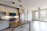 This contemporary single-floor residence showcases a thoughtfully designed open-concept layout that seamlessly connects the kitchen and living areas. The kitchen is a standout feature, equipped with stainless steel appliances including a gas range, built-in microwave, dishwasher, and a French door refrigerator. Flat-panel cabinetry in a warm wood-tone and soft gray finish lines the walls, complemented by an intricate geometric tile backsplash and dark quartz countertops. A mobile kitchen island provides additional prep space and flexibility. Recessed and track lighting illuminate the kitchen, while wood-look plank flooring extends throughout the main living areas, adding warmth and continuity.

The living area is bright and airy, benefiting from floor-to-ceiling windows and a glass door that opens to a private balcony with outdoor views. A ceiling fan adds both function and style to the space.

The bedroom continues the theme of natural light, featuring large windows with balcony access, soft carpeting underfoot, and a ceiling fan overhead, creating a comfortable and tranquil retreat.

The bathroom is finished to a high standard, presenting a dual-sink floating vanity with dark quartz countertops, wood-grain cabinetry, matte black fixtures, and LED-backlit mirrors. Large-format floor tiles complete the clean, modern aesthetic throughout.