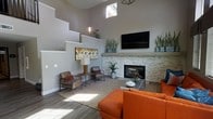 This contemporary lounge space makes an immediate impression with its dramatic, soaring ceilings that amplify the sense of openness and volume. Neutral gray walls serve as a clean backdrop, allowing the design elements to take center stage. Natural light filters in through clerestory windows positioned high on the walls, casting warm sunlight across the hardwood-style flooring below.

The focal point of the space is a beautifully crafted stacked stone fireplace, topped with a sleek concrete-style mantel and flanked by lush greenery in modern planters. A wall-mounted flat-screen television sits above the mantel, integrating entertainment seamlessly into the design. The seating arrangement features bold burnt-orange upholstered furniture paired with rich brown leather accent chairs, creating a vibrant yet cohesive color palette. A patterned area rug anchors the seating area, adding texture and warmth underfoot.

Architectural interest is provided by a striking stepped wall detail that adds dimension and visual depth to the room. Thoughtful touches such as wall sconces, curated artwork, and carefully placed indoor plants contribute to the polished, yet approachable ambiance. Overall, this space blends modern design with comfortable functionality, making it an ideal setting for relaxation or social gathering.