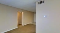 This single-floor space presents a straightforward, functional layout with a calm, neutral aesthetic throughout. The main living area is finished with wall-to-wall carpeting in a warm beige tone, complementing the similarly toned painted walls that create a cohesive and understated interior palette. A textured popcorn ceiling adds a classic touch overhead, while a ceiling-mounted HVAC vent ensures climate control throughout the space. A thermostat is conveniently positioned on a nearby wall for easy access. The room opens toward a doorway leading to an additional area, suggesting a connected layout that allows for a natural flow between spaces. A second door, likely a closet, is visible along the far wall, offering built-in storage. The space is well-lit by overhead lighting visible through the connecting doorway, contributing to a bright and welcoming atmosphere. Overall, the unit offers a clean, move-in-ready environment with practical finishes suited for everyday living.
