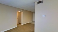 This single-floor space presents a straightforward, functional layout with a calm, neutral aesthetic throughout. The main living area is finished with wall-to-wall carpeting in a warm beige tone, complementing the similarly toned painted walls that create a cohesive and understated interior palette. A textured popcorn ceiling adds a classic touch overhead, while a ceiling-mounted HVAC vent ensures climate control throughout the space. A thermostat is conveniently positioned on a nearby wall for easy access. The room opens toward a doorway leading to an additional area, suggesting a connected layout that allows for a natural flow between spaces. A second door, likely a closet, is visible along the far wall, offering built-in storage. The space is well-lit by overhead lighting visible through the connecting doorway, contributing to a bright and welcoming atmosphere. Overall, the unit offers a clean, move-in-ready environment with practical finishes suited for everyday living.