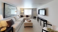 This single-floor residence is thoughtfully laid out to maximize functionality across its key living spaces. The living room serves as the heart of the home, featuring a large, cream-toned sectional sofa paired with a matching ottoman, soft carpeting, and a wall-mounted flat-screen television. A large framed map print anchors the seating area, while a window with grey curtains and an in-unit air conditioning unit allows for natural light and climate control. The space is styled with a cohesive neutral palette, complemented by dark-accented furniture pieces.

Adjacent to the living room, the dining area accommodates a round wooden dining table surrounded by cross-back chairs, centered beneath a ceiling fan with a light fixture. A compact bar cart and a decorative mirror add character to the corner of the room.

The galley-style kitchen is efficiently designed with white cabinetry, light countertops, a coil-burner electric range, and a refrigerator. Tile flooring and under-cabinet ventilation complete the functional layout.

The bathroom features a full-sized soaking tub with a shower and curtain, tile flooring, and clean white fixtures, maintaining the home's consistent neutral and understated aesthetic throughout.