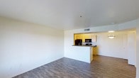 This single-floor space offers a clean, open-concept layout that seamlessly integrates the living area and kitchen into one cohesive environment. The living area is bright and airy, featuring wood-look hardwood-style flooring that runs throughout, lending warmth and continuity to the space. Smooth white walls and a flat white ceiling contribute to a sense of openness and light.

The kitchen is defined by a peninsula-style counter with a dark laminate countertop, which serves as a natural divider between the living and cooking areas while maintaining an open sightline. The cabinetry is finished in a warm honey-toned wood, providing a welcoming contrast to the neutral walls. Black appliances, including a microwave and range, are integrated into the kitchen layout, offering a modern and functional cooking space. A ceiling-mounted HVAC vent ensures consistent climate control throughout the unit.

The overall ambiance of the space is clean, minimalist, and move-in ready, with a straightforward design that prioritizes functionality and flow. The open floor plan makes efficient use of the available space, allowing natural light to reach the living area and creating a versatile environment suitable for a variety of living arrangements.