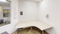 Located at 651 W 6th St in Gilroy, California, this single-floor community facility offers a well-organized and functional layout across a generously sized floor plan. The space is designed to accommodate a variety of activities and gatherings, with distinct zones that serve different purposes.

The main hall forms the heart of the facility, featuring rows of modular tables and chairs arranged to support classroom-style programming or group workshops. Suspended drop-tile ceilings with integrated fluorescent lighting ensure consistent illumination throughout, while vertical blind-covered windows invite natural light and views of the surrounding greenery.

A dedicated meeting or presentation room is furnished with rows of stackable chairs facing large arched windows, making it well-suited for assemblies, lectures, or community presentations. Adjacent to the main hall is a comfortable lounge area appointed with upholstered armchairs, a central coffee table, a wall-mounted television, and an American flag — providing a welcoming space for informal gatherings.

A smaller, enclosed private workspace features an L-shaped desk and posted informational materials, offering a quiet area for individual use. A reception corridor connects the key areas, complete with a brochure display rack and a windowed service partition. Carpeted flooring runs throughout the space, contributing to a warm and accessible atmosphere suitable for community-oriented programming.