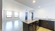 This single-floor space showcases a thoughtfully designed open-concept layout that seamlessly connects the kitchen and living areas. The kitchen is a standout feature, equipped with dark espresso cabinetry, granite countertops with subtle patterning, a peninsula that provides additional prep space and casual seating, a built-in electric cooktop, and stainless steel appliances. A mosaic tile backsplash adds a refined decorative touch to the kitchen wall. The polished concrete floors extend throughout the space, lending an industrial-modern aesthetic that is both durable and visually striking. Recessed lighting is installed throughout, providing consistent and adjustable illumination across the open floor plan. The living area is bright and open, benefiting from a large window that invites natural light and views of the surrounding environment. The crisp white walls and high ceilings contribute to a clean, airy atmosphere. Two doors visible toward the rear of the space lead to additional rooms, suggesting private quarters separate from the main living and kitchen area. Overall, the space combines contemporary finishes with functional design for a polished, modern feel.