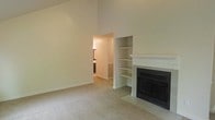 This single-floor residence offers a thoughtfully designed layout that balances comfort and functionality. The main living area immediately impresses with its dramatically high ceilings, which lend a sense of openness and grandeur to the space. Neutral tones throughout — from the soft beige carpet underfoot to the crisp white walls — create a clean, welcoming ambiance that is easy to personalize.

A standout feature of the living room is the classic fireplace, framed by a white mantel and finished with dark tile surround, making it a natural focal point for the room. Adjacent to the fireplace, a set of built-in open shelves provides practical display and storage space while adding architectural character to the wall.

An open passageway connects the living area to the rest of the home, offering a glimpse into a hallway that leads toward additional rooms, including what appears to be a kitchen or utility space with warm cabinetry visible in the background. This seamless flow between spaces enhances the overall sense of connectivity within the home.

The wall-to-wall carpeting adds warmth and comfort throughout the living area, while the generous ceiling height and open floor plan ensure the space feels expansive and inviting. Overall, this residence presents a clean, move-in-ready environment with timeless design details.