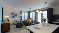 This contemporary single-floor residence offers a thoughtfully designed open-concept layout that seamlessly connects its key living spaces. A central hallway serves as the spine of the unit, providing clear sightlines to both the kitchen and bedroom, while built-in open shelving along one wall offers practical display and storage space.

The kitchen features a dark, striking island with a light-colored countertop, complemented by pendant lighting overhead that adds a modern touch. It flows directly into the living area, which is bathed in natural light courtesy of a generous bank of large windows dressed with patterned drapes and horizontal blinds. The living room is anchored by a comfortable seating arrangement and finished with wood-look flooring that extends throughout the space, lending warmth and visual continuity.

The bedroom, visible from the hallway through a dark-framed door, enjoys its own bright windows and is finished with the same cohesive flooring and neutral color palette found throughout the residence. Recessed lighting in the hallway and living areas ensures consistent, even illumination. The overall ambiance is clean, modern, and inviting, with a neutral color scheme accented by warm metallic and dark wood tones that give the space a polished, contemporary character.