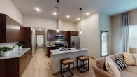 This well-appointed single-floor residence offers a thoughtfully designed open-concept layout with a seamless flow between the kitchen, dining, and living areas. The kitchen serves as the centerpiece of the home, showcasing rich dark wood cabinetry that reaches toward the impressively high ceilings, complemented by granite countertops and a full suite of stainless steel appliances including a gas range, built-in microwave, and a French door refrigerator. A spacious kitchen island with a granite surface and undermount sink provides additional prep space and casual seating with industrial-style bar stools. Elegant globe pendant lights hang above the island, adding a refined touch to the space. Light wood-tone flooring runs throughout the main living areas, creating a warm and cohesive aesthetic. The living room features large windows dressed with patterned curtains, allowing ample natural light to fill the room. The bedroom offers plush carpeting, a large window with blinds, and soaring ceiling height that adds to the sense of openness. The bathroom is finished with a granite-topped dark wood vanity, an illuminated mirror, and a full tub-shower combination. Recessed lighting throughout ensures each room is well-lit and inviting.