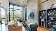 Located at 320 S Walker St in Cary, NC, this single-floor space makes a bold architectural statement with its soaring double-height ceilings and sophisticated, contemporary design. The main lounge area centers around a dramatic floor-to-ceiling natural stone accent wall, housing a linear fireplace and mounted display screen flanked by built-in navy shelving. Towering steel-frame windows flood the space with natural light and offer views of the surrounding greenery, while a sculptural floral chandelier adds a refined finishing touch overhead. Warm hardwood flooring runs throughout, tying each area together cohesively.

The kitchen is a standout, outfitted with sage green cabinetry, a gray subway tile backsplash, brass and matte black fixtures, stainless steel appliances, and a generous waterfall-edge quartz island. Pendant lighting with gold accents adds warmth to the space. A secondary lounge area is accessible through dramatic frosted-glass barn doors with black metal framing, offering a more intimate setting with cognac leather seating and a decorative living wall of curated botanicals and art objects. A long communal table and additional seating provide a versatile workspace or gathering area, completing this well-appointed and design-forward interior.