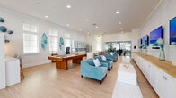 Located at 2130 Mayport Rd in Jacksonville, FL, this single-floor amenity clubroom offers a generously sized, open-plan gathering space designed with a vibrant coastal aesthetic. Light hardwood flooring flows throughout, complementing the crisp white walls and bright, recessed lighting overhead. A standout feature wall showcases a row of flat-screen TVs mounted above a built-in white cabinetry unit with a warm wood countertop, creating a dedicated entertainment zone framed by nautical-inspired décor. Opposite this wall, a full kitchen area provides a large white island with bar seating, glass-front upper cabinets, a decorative tile backsplash, stainless steel appliances, and stylish pendant lighting — ideal for social gatherings and events. A billiards table anchors the central recreation zone, surrounded by plush teal accent chairs and ocean-themed wall art, including surfboard sculptures and resin pieces that reinforce the coastal atmosphere. A dining area near floor-to-ceiling windows and glass doors floods the space with abundant natural light while offering direct views of palm trees and an outdoor pool area beyond. An adjacent glass-walled fitness center is visible from the main space, adding functional versatility. The overall ambiance is bright, welcoming, and distinctly Florida coastal in character.