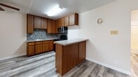 This single-floor unit offers a practical and cohesive layout with consistent light gray wood-look flooring running throughout, tying each area together seamlessly. The kitchen is a standout feature, equipped with warm oak cabinetry that extends to the ceiling for generous storage, a mosaic tile backsplash in neutral tones, a gas range, stainless steel over-the-range microwave, and a double sink with ample counter space. A peninsula with a smooth countertop extends into the living area, creating a natural breakfast bar and open sightlines between the kitchen and main living space. The open layout of the kitchen and living area is visible in the overhead dollhouse view, showcasing the efficient use of space. The bathroom features a wood-toned vanity with a white countertop, a mirrored medicine cabinet, and a separate water closet housing a shower-tub combination with a curtain rod and toilet — all finished in clean, neutral tones. The bedroom is bright and airy, with soft beige walls, a textured ceiling, sliding double closet doors offering built-in storage, and a window dressed with vertical blinds that allows natural light to filter in. Throughout the space, white baseboards and trim provide a clean, finished look.