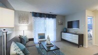 This single-floor space showcases a thoughtfully designed living area centered around a calming teal and neutral color palette. The room is anchored by a plush gray sofa adorned with teal accent pillows, complemented by a patterned accent chair that adds visual interest without overwhelming the space. A sleek, dark coffee table with a chrome frame serves as the focal point of the seating arrangement, styled with decorative objects that echo the room's color scheme.

Natural light fills the space through a large window dressed with layered curtains — sheer patterned panels beneath teal grommet-top drapes — creating an airy, welcoming atmosphere. The window treatment allows diffused light to brighten the neutral-toned walls and light carpet flooring throughout.

A modern white media console lines one wall, providing clean storage while supporting a flat-screen television above. Decorative wall art in metallic finishes flanks both sides of the room, adding texture and a touch of sophistication. The textured ceiling and soft, neutral tones throughout contribute to a comfortable and stylish ambiance suited for everyday living.