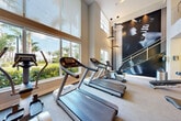 Located at 1402 S Dixie Hwy in Lantana, Florida, this well-appointed two-story fitness center offers a comprehensive workout experience across two generously sized floors connected by a sleek staircase with cable railings. The ground floor serves as the primary exercise area, featuring an extensive selection of strength training machines, free weights, a power rack with benches, and a full row of cardio equipment including treadmills, ellipticals, and rowing machines. Oversized windows flood the space with natural light and frame lush tropical greenery outside, creating an energizing atmosphere. A striking large-scale artistic mural serves as a bold focal point, complementing the modern aesthetic defined by neutral tones, carpeted flooring, and recessed linear lighting. Structural columns punctuate the open floor plan, adding architectural definition without interrupting the flow of the space. The upper floor houses a dedicated studio room finished with hardwood-style flooring and floor-to-ceiling mirrors along the walls, ideal for cycling, rowing, and group fitness activities. Additional equipment and open workout space on the upper level overlook the main floor below. A utility room and support spaces round out the facility's functional offerings.