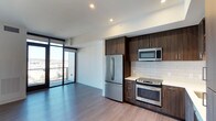 Located at 300 14th Street, Unit 1305 in San Diego, this contemporary single-floor residence offers a thoughtfully designed open-concept layout. The living and kitchen areas flow seamlessly together, anchored by rich dark wood cabinetry that extends to the ceiling for maximum storage. The kitchen is outfitted with stainless steel appliances, including a French door refrigerator, a range with oven, and an over-the-range microwave. A classic white subway tile backsplash and light countertops provide a clean, modern contrast to the cabinetry. Wide-plank hardwood-style flooring runs throughout, adding warmth and continuity to the space. Recessed lighting illuminates the interior, complementing the abundant natural light that pours in through expansive floor-to-ceiling windows along the exterior wall. A sliding glass door opens to a private balcony, offering elevated views of the surrounding San Diego cityscape. The open living area benefits directly from this wall of glass, creating a bright and airy atmosphere. The overall aesthetic is modern and polished, with a neutral palette and high-quality finishes that reflect contemporary urban living at its finest.