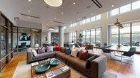 This contemporary single-floor community lounge is designed to encourage social interaction and relaxed gathering. The open-plan layout is anchored by soaring ceilings with clerestory windows that flood the interior with natural light, complemented by warm hardwood flooring throughout.

The main lounge area features generous leather and upholstered sofas adorned with colorful accent pillows, centered around a wooden coffee table. Adjacent to this is a vibrant seating nook with patterned armchairs set against a wall of large picture windows, offering views of the surrounding greenery.

A dedicated game and casual dining zone includes a shuffleboard table alongside white dining tables paired with colorful modern chairs in teal and green, with a wall-mounted flat-screen television for entertainment. Lush potted tropical plants are strategically placed throughout, adding a natural, refreshing element to the interior.

A kitchenette area is integrated into the space, featuring appliances and counter seating beneath stylish pendant lighting. Glass-partitioned rooms along the perimeter provide more private spaces for focused work or meetings, visible through steel-framed glazing. Cylindrical architectural columns punctuate the open floor plan, reinforcing the building's structural character. The overall ambiance is vibrant, welcoming, and thoughtfully curated for both relaxation and productivity.