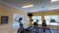 This single-floor fitness center is designed to provide a functional and comfortable workout environment. The room features warm yellow walls accented by crisp white crown molding and baseboards, lending the space a clean, polished look. Flush-mounted fluorescent light panels illuminate the room evenly, complemented by natural light streaming through multiple large windows fitted with white horizontal blinds. A wall-mounted flat-screen television is positioned for easy viewing during workouts, adding to the amenity's convenience. The space is outfitted with a variety of cardio and strength training equipment, including an elliptical machine, a treadmill, stationary bikes, and additional resistance machines, offering a well-rounded selection for users of varying fitness levels. Dark flooring provides a practical contrast to the light-toned walls and ceilings, while a small shelf beneath the mounted television offers storage for personal items. The overall ambiance is bright, clean, and purposefully organized, making this fitness center a practical and inviting space for daily exercise routines.