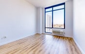 Located in Woodside, Queens, this thoughtfully designed studio unit offers a clean, modern aesthetic across a single open floor plan. Light-toned hardwood flooring runs continuously throughout, lending warmth and cohesion to the space.

The living area is bright and airy, anchored by a striking floor-to-ceiling window with bold black framing that frames panoramic views of the Woodside and Queens skyline, flooding the room with natural light. A built-in HVAC unit ensures year-round comfort.

The kitchen is seamlessly integrated into the layout, featuring sleek white cabinetry that extends to the ceiling for ample storage. Stainless steel appliances — including a gas range and a full-size bottom-freezer refrigerator — pair with white countertops and a matte black sink fixture for a polished, contemporary look. A dedicated closet with built-in rods is conveniently positioned adjacent to the kitchen.

The hallway connects the living space to the bathroom, which is finished with large-format neutral tiles on both the floors and walls. A light wood vanity with a modern vessel sink, matte black fixtures, and a recessed medicine cabinet complete the refined, spa-like atmosphere. White walls and recessed lighting throughout contribute to the unit's clean, minimalist character.