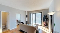 This single-floor space features a well-lit living area anchored by large sliding glass doors that open to a balcony, allowing abundant natural light to fill the room and offering views of the surrounding neighborhood. The living area is finished with light hardwood flooring, which adds warmth throughout the space, and features a textured ceiling. A tufted sofa and glass coffee table furnish the main area, creating a comfortable and functional gathering space. A structural column with a decorative tile accent adds a subtle architectural detail near the sliding doors. Adjacent to the living area, a bedroom is visible through an open doorway, also benefiting from natural light through its own window. The overall layout flows openly between spaces, with neutral gray walls providing a clean, versatile backdrop throughout. A coat and storage area is situated near the balcony entrance, offering practical organization at the entry point. The combination of natural light, hardwood floors, and access to outdoor space gives this unit a bright and livable character well-suited for everyday comfort.