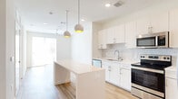 This thoughtfully designed one-bedroom, one-bathroom unit offers a clean, modern aesthetic across a single floor. The open-concept kitchen serves as the centerpiece of the living area, featuring crisp white cabinetry, a subtle tile backsplash, and a full suite of stainless steel appliances including a French door refrigerator, electric range, microwave, and dishwasher. A white quartz center island provides additional prep space and casual seating, while two globe pendant lights with warm gold interiors add a stylish focal point overhead. Warm-toned hardwood-style flooring flows throughout the main living areas, creating a cohesive and inviting atmosphere. Natural light enters through sliding glass doors in the living area, enhancing the bright, airy feel of the space. The bedroom is comfortably sized with plush carpeting, a large window offering neighborhood views, and recessed lighting. A hallway connects the bedroom to the full bathroom, which is finished with a white vanity and wood-tone flooring, and leads directly to a dedicated walk-in closet with built-in shelving. In-unit stacked laundry is also conveniently accessible. The overall design prioritizes clean lines, neutral tones, and functional layout throughout.