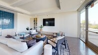 This contemporary single-floor amenity space is thoughtfully designed to blend comfort, functionality, and modern aesthetics. The interior is anchored by polished terrazzo-style concrete flooring that extends throughout, lending a clean and sophisticated foundation to the entire space.

The main lounge area is a generously proportioned gathering space featuring a coffered ceiling with wood-paneled insets and recessed lighting that adds warmth and architectural interest overhead. A large wall-mounted flat-screen television serves as a focal point, flanked by a sleek built-in media console and a floor-to-ceiling open shelving unit finished in light wood tones. Comfortable seating arrangements — including plush sofas and accent armchairs in neutral tones — are grouped around a round glass coffee table atop a layered area rug, creating an inviting setting for socializing or relaxing. Large windows on the perimeter allow abundant natural light to fill the room and offer views of the surrounding outdoor area.

Adjacent to the lounge is a bright dining area enclosed by expansive floor-to-ceiling glass walls and sliding glass doors on multiple sides. This sun-drenched space accommodates several dining tables with woven chairs and opens directly onto a landscaped outdoor courtyard, where palm trees, lawn seating, and a pergola structure are visible. The seamless indoor-outdoor connection makes this area ideal for casual gatherings and community events.