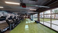 Located at 408 N Terry Ave in Orlando, FL, this gym is a comprehensively outfitted fitness facility designed to support a wide range of training styles. The space is defined by an industrial-chic aesthetic, featuring exposed ceilings with visible ductwork and structural beams, illuminated by overhead linear LED lighting throughout.

A standout feature is the row of large glass-panel garage doors along one side of the facility, which flood the interior with natural light and offer open views of a lush outdoor courtyard lined with palm trees. The artificial turf flooring that runs through a significant portion of the gym reinforces an energetic, athletic atmosphere ideal for functional training and agility work.

The cardio zone is well-stocked with StairMaster steppers, ellipticals, rowing machines, and spin bikes. The strength training area houses a full rack of dumbbells, barbells, weight plates, benches, and a cable/multi-use rig equipped with a punching bag and TRX suspension trainers. Medicine balls, kettlebells, and plyo boxes are neatly organized for functional fitness workouts.

Bold, large-scale graphic murals adorn the walls, adding a dynamic visual energy to the space. A mirrored wall section in the strength area enhances the sense of openness. The facility also includes a dedicated entry and restroom access, making it a complete, self-contained fitness destination.