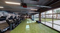 Located at 408 N Terry Ave in Orlando, FL, this gym is a comprehensively outfitted fitness facility designed to support a wide range of training styles. The space is defined by an industrial-chic aesthetic, featuring exposed ceilings with visible ductwork and structural beams, illuminated by overhead linear LED lighting throughout.

A standout feature is the row of large glass-panel garage doors along one side of the facility, which flood the interior with natural light and offer open views of a lush outdoor courtyard lined with palm trees. The artificial turf flooring that runs through a significant portion of the gym reinforces an energetic, athletic atmosphere ideal for functional training and agility work.

The cardio zone is well-stocked with StairMaster steppers, ellipticals, rowing machines, and spin bikes. The strength training area houses a full rack of dumbbells, barbells, weight plates, benches, and a cable/multi-use rig equipped with a punching bag and TRX suspension trainers. Medicine balls, kettlebells, and plyo boxes are neatly organized for functional fitness workouts.

Bold, large-scale graphic murals adorn the walls, adding a dynamic visual energy to the space. A mirrored wall section in the strength area enhances the sense of openness. The facility also includes a dedicated entry and restroom access, making it a complete, self-contained fitness destination.