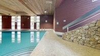 Located at 252-254 Gorge Rd E in Victoria, British Columbia, this indoor aquatic facility offers a generously sized, single-level pool space designed for both recreation and fitness. The centrepiece is a large, clear-water swimming pool with a classic mosaic tile surround, pool entry railings, and underwater lighting that gives the water a vivid turquoise appearance. The architectural character of the space is defined by warm, horizontal cedar-toned wood panelling that lines the lower half of the walls, complemented by crisp white upper walls and a distinctive natural wood plank ceiling that lends a welcoming, chalet-like warmth to the interior. A striking dry-stacked stone accent wall adds a rugged, organic texture near the staircase entry, providing visual contrast to the polished tile deck. Tall, floor-to-ceiling windows along one side of the facility flood the pool area with natural light and offer views of surrounding greenery. The space is equipped with HVAC units to maintain a comfortable climate year-round. A staircase provides access to an upper-level viewing or amenity area. Safety equipment, including a life ring, is visibly stationed poolside. The overall ambiance is bright, clean, and functional, making this a well-appointed indoor aquatic facility suitable for lap swimming and leisure use.