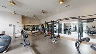 This fitness center offers a generously proportioned, single-level workout space designed to accommodate a wide variety of training styles. The layout is thoughtfully organized, with distinct zones for strength training and cardiovascular exercise. The strength training area features a multi-station power rack with pull-up bars, cable machines, free weights, barbells with plate storage, and adjustable benches — providing ample options for resistance-based workouts. Large mirrors line the walls throughout, visually expanding the space and allowing users to monitor their form. The cardio zone, positioned along a wall of large, grid-paned windows, houses treadmills, elliptical machines, stair climbers, and rowing machines. Natural light flows generously through these oversized windows, creating a bright and energizing atmosphere. The flooring throughout is durable rubber, appropriate for heavy use and equipment placement. Multiple ceiling fans are mounted overhead, contributing to air circulation and comfort. Recessed lighting supplements natural light during all hours. A vending area is visible in the background, adding convenience for users. Overall, the space presents a clean, functional, and well-maintained fitness environment.