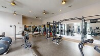 This fitness center offers a generously proportioned, single-level workout space designed to accommodate a wide variety of training styles. The layout is thoughtfully organized, with distinct zones for strength training and cardiovascular exercise. The strength training area features a multi-station power rack with pull-up bars, cable machines, free weights, barbells with plate storage, and adjustable benches — providing ample options for resistance-based workouts. Large mirrors line the walls throughout, visually expanding the space and allowing users to monitor their form. The cardio zone, positioned along a wall of large, grid-paned windows, houses treadmills, elliptical machines, stair climbers, and rowing machines. Natural light flows generously through these oversized windows, creating a bright and energizing atmosphere. The flooring throughout is durable rubber, appropriate for heavy use and equipment placement. Multiple ceiling fans are mounted overhead, contributing to air circulation and comfort. Recessed lighting supplements natural light during all hours. A vending area is visible in the background, adding convenience for users. Overall, the space presents a clean, functional, and well-maintained fitness environment.
