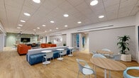 Located at 325 N 15th St in Philadelphia, PA, this thoughtfully designed commercial lounge space occupies a single floor and offers a generous, open-plan layout tailored for collaboration and comfort. The interior is organized into several distinct seating zones that flow seamlessly into one another, all unified by warm light wood flooring and a clean white ceiling fitted with recessed lighting and striking geometric pendant fixtures.

A casual lounge area anchors one section of the space, featuring a large sectional sofa in teal, complemented by chartreuse accent chairs and a round coffee table, all set atop a patterned area rug. A wall-mounted flat-screen TV and sleek media console complete this inviting retreat. Adjacent to this, a vibrant social zone showcases bold orange sofas arranged beneath framed wall art and surrounded by lush potted greenery.

Café-style seating areas with round bistro tables and contemporary chairs provide flexible informal gathering spots throughout. Tall arched windows framed by exposed brick columns allow natural light to filter in, adding warmth and character. A reception or service desk with wood and glass detailing anchors one end of the space, while glass-partitioned enclosures offer semi-private areas. The overall ambiance is modern, energetic, and welcoming—ideal for a professional yet comfortable communal environment.
