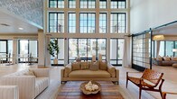 This elegantly designed amenity lounge is a standout communal space defined by its dramatic double-height ceilings and an expansive wall of black-framed, floor-to-ceiling windows that flood the interior with natural light and offer unobstructed views of the outdoor pool and courtyard area. The main lounge area centers around a plush neutral-toned sofa arrangement atop a layered area rug, complemented by woven accents and coastal-inspired décor that establish a relaxed yet refined aesthetic throughout.

A striking patterned tile fireplace with a natural wood mantel and wall-mounted television serves as a focal point on one end of the lounge, flanked by sleek barn-style sliding doors with black steel grid detailing that elegantly divide the space. Light wide-plank hardwood flooring extends seamlessly across the entire area, enhancing the open, airy feel.

The space also includes a well-appointed kitchen and café bar area featuring a large white quartz island with rattan bar seating, a dedicated coffee and refreshment station with open shelving, a stainless steel refrigerator, and shiplap wall accents. A separate casual dining nook with bistro tables sits adjacent to sliding glass doors that open directly to the outdoor terrace, further blurring the line between indoor and outdoor living. Wall sconces, pendant lighting, and a statement chandelier complete the warm, welcoming ambiance.