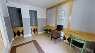 This single-floor space functions as a shared business center or common area workroom, thoughtfully designed to support productivity in a visually dynamic environment. The room is defined by its bold yellow trim and panel framing along the walls, which contrast sharply with the two-tone gray and white wall treatment, creating a lively and energetic atmosphere.

The workstation area features a continuous yellow-trimmed desk surface that runs along the wall, accommodating a desktop computer setup and providing ample workspace. Seating is eclectic and intentional, mixing a woven rattan armchair with vibrant green chairs and stools, lending the space a casual yet functional character. Decorative diamond-patterned storage cabinets in yellow, purple, and dark tones add a playful geometric design element while offering practical storage.

The flooring consists of neutral light beige tiles accented by a bordered area rug inset, grounding the room with a clean and polished finish. A recessed alcove with additional cabinetry provides supplementary storage or utility space. Framed botanical artwork on the wall adds a subtle decorative touch. Overall, the space balances functionality with a distinctive, colorful aesthetic that makes it both practical and visually engaging.