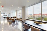Located at 325 15th Street in Jersey City, NJ, this expansive single-floor amenity space offers a refined and contemporary environment designed for comfort and social engagement. The layout spans a generous footprint and is organized into distinct yet flowing zones, each thoughtfully curated with modern furnishings and design-forward details.

The living area features a warm caramel leather sofa and a pair of cream upholstered accent chairs arranged around a marble coffee table, all set against cool gray walls adorned with vibrant abstract artwork. Polished concrete and marbled epoxy flooring run throughout, reflecting light and lending the space a sleek, upscale character.

A central lounge zone includes two channel-tufted sofas flanking a large wall-mounted television, illuminated by a striking circular ring chandelier suspended from the ceiling. Recessed lighting complements the ambient glow throughout.

The dining and gathering area is bathed in natural light from expansive floor-to-ceiling windows that frame sweeping views of the surrounding Jersey City skyline and outdoor terrace. A large oval conference-style dining table with modern chairs anchors this zone, accompanied by a built-in bar counter with tall seating. Track and pendant lighting add layered illumination.

Together, these areas create a polished, welcoming environment ideal for both relaxed gatherings and productive meetings.