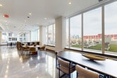 Located at 325 15th Street in Jersey City, NJ, this expansive single-floor amenity space offers a refined and contemporary environment designed for comfort and social engagement. The layout spans a generous footprint and is organized into distinct yet flowing zones, each thoughtfully curated with modern furnishings and design-forward details.

The living area features a warm caramel leather sofa and a pair of cream upholstered accent chairs arranged around a marble coffee table, all set against cool gray walls adorned with vibrant abstract artwork. Polished concrete and marbled epoxy flooring run throughout, reflecting light and lending the space a sleek, upscale character.

A central lounge zone includes two channel-tufted sofas flanking a large wall-mounted television, illuminated by a striking circular ring chandelier suspended from the ceiling. Recessed lighting complements the ambient glow throughout.

The dining and gathering area is bathed in natural light from expansive floor-to-ceiling windows that frame sweeping views of the surrounding Jersey City skyline and outdoor terrace. A large oval conference-style dining table with modern chairs anchors this zone, accompanied by a built-in bar counter with tall seating. Track and pendant lighting add layered illumination.

Together, these areas create a polished, welcoming environment ideal for both relaxed gatherings and productive meetings.