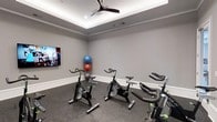 This single-floor fitness studio is thoughtfully designed to provide a focused and functional workout environment. The spin cycle room serves as a centerpiece of the space, featuring multiple stationary bikes arranged for group or individual cycling sessions. A wall-mounted flat-screen display faces the bikes, enabling guided workout streams or entertainment during rides. A dedicated exercise ball rack holds multiple stability balls of varying sizes, adding versatility for strength and core training. The room is finished with durable rubber flooring, ideal for high-impact activity and equipment stability. Neutral gray walls contribute to a clean, modern aesthetic throughout, while crisp white crown molding and baseboards add a polished architectural touch. A recessed tray ceiling with integrated ambient lighting creates a bright and energizing atmosphere, complemented by a ceiling fan that ensures comfortable air circulation during intense workouts. The space is equipped with ventilation fixtures to maintain a fresh environment. Overall, the studio balances practical fitness functionality with refined interior finishes, making it a well-rounded facility suited for cardio and cross-training workouts.