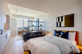 Located at 10 E Reed St in San Jose, California, this thoughtfully designed one-bedroom, two-bathroom residence offers a modern aesthetic with clean architectural lines throughout. Light-toned hardwood flooring flows continuously across the single-level layout, creating a cohesive and bright atmosphere.

The kitchen is a standout feature, showcasing sleek flat-panel cabinetry in white and warm wood tones, a white tile backsplash, integrated appliances including a built-in oven and induction cooktop, and a stainless steel sink. Floor-to-ceiling windows adjacent to the kitchen and living area flood the space with natural light and provide expansive city views of San Jose.

The bedroom is a well-proportioned retreat featuring a full wall of built-in wardrobes in soft grey with warm wood handles, flanking a navy blue open shelving unit with integrated drawers — a functional and stylish storage solution. Recessed lighting adds a clean finish overhead.

The two bathrooms are finished to a high standard. One features a full walk-in shower clad in floor-to-ceiling sage green mosaic tiles with chrome fixtures. The second bathroom includes a floating white vanity with an illuminated mirror, green mosaic tile flooring, and curated wall art, lending it a refined, gallery-like feel. A dedicated entrance completes this efficiently designed urban residence.