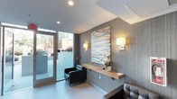 This single-floor space presents a thoughtfully designed lobby or reception area that combines modern aesthetics with functional elements. The interior is anchored by textured gray wall paneling that adds depth and sophistication, complemented by clean white ceilings with recessed lighting. A striking large-scale abstract artwork featuring diagonal lines in muted blues, oranges, and whites serves as a central focal point on the main wall. Flanking the artwork, warm-toned wall sconces provide ambient lighting that enhances the welcoming atmosphere. A minimalist floating wood shelf runs along the feature wall, styled with a white orchid arrangement, adding a natural, elegant touch. Seating is provided by sleek black leather chairs, offering a professional yet comfortable waiting area. Large floor-to-ceiling glass panels and a glass entry door flood the space with natural light and provide views of the exterior surroundings, creating a seamless connection between indoors and outdoors. Light-toned large-format floor tiles contribute to the bright, open feel of the space. Overall, the lobby conveys a polished, contemporary character suited to a professional or commercial environment.
