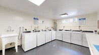 Located at 18540 Plummer Street in the Northridge neighborhood of Los Angeles, this shared laundry facility is a well-organized and functional space designed to serve multiple users efficiently. The room is illuminated by a combination of skylights and flush-mounted ceiling light panels, creating a bright and clean environment. The light-colored walls and dark speckled flooring give the space a tidy, utilitarian aesthetic.

The laundry room is equipped with a row of top-loading washing machines lined along one wall, paired with a corresponding row of front-loading dryers, providing ample capacity for shared use. Each machine features digital controls for ease of operation. Plumbing hookups are clearly visible along the walls, and posted signage — including laundry room rules and machine issue notices — keeps users well-informed.

A wall-mounted folding counter runs along one side of the room, offering a convenient surface for sorting and folding laundry. A decorative "Laundry Room" sign adds a welcoming touch to the space. The room is accessed through a sturdy metal security door, and a small waste bin is provided for tidiness. A water heater is also present, supporting the facility's operational needs. Overall, the space is practical, well-lit, and thoughtfully arranged for shared community use.