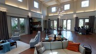 This elegant space makes an immediate impression with its dramatic double-height ceilings and a wall of clerestory windows that flood the interior with natural light. A striking cascading chandelier with spherical glass accents serves as a focal point, anchoring the expansive open-plan layout beneath a coffered ceiling with integrated uplighting.

The main lounge area is thoughtfully furnished with neutral-toned sofas and accent chairs arranged around a sleek rectangular coffee table, accented with decorative teal vases and lush greenery. Rich, dark hardwood flooring runs throughout, providing a warm contrast to the light walls and modern décor.

Beyond the seating area, a casual dining zone features dark bistro-style tables and chairs, positioned near floor-to-ceiling glass doors that open to lush outdoor greenery, seamlessly blending indoor and outdoor environments. To one side, a well-appointed bar or refreshment counter with dark cabinetry offers a convenient amenity space.

The overall ambiance is polished yet inviting, with a neutral palette of taupe, grey, and warm brown tones complemented by pops of teal and orange. Large glass doors on multiple walls enhance the sense of openness and connectivity to the outdoors, making this a versatile and sophisticated communal space.